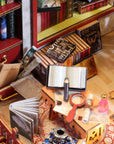 A miniature dollhouse interior scene featuring a wooden desk cluttered with tiny books, an open book, a magnifying glass, ink bottle, and other study accessories, surrounded by miniature bookshelves and furniture.