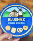 A blue tin with a cookie design and the text 'BLUSHIEZ Butter Cookies' on it, held by a hand. The tin contains various shaped cookies and a spool of thread.