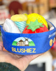 A blue tin with a cookie design and the text 'BLUSHIEZ Butter Cookies' on it, held by a hand. The tin is opend and shows various shaped cookies and a spool of thread.