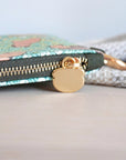A zipper view of the multicolored coin purse with a brass zipper and a metal key ring, featuring a pattern of capybaras, flowers, and leaves with a fabric accent in the background