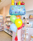 A balloon arrangement featuring a pencil pouch balloon, with colorful latex balloons, a handmade tassel, and a personalized grade tag, displayed in a store setting.