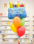 A balloon arrangement featuring a pencil pouch balloon, with colorful latex balloons, a handmade tassel, and a personalized grade tag, displayed in a store setting with card shelves