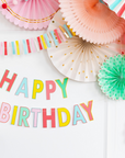 A birthday banner with the words 'HAPPY BIRTHDAY' displayed in colorful letters against a wall with other paper fan decor