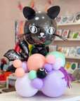 A decorative balloon arrangement shaped like a black cat head on a cluster of pastel balloons, including lavender, peach, mint green, pink, and purple, photographed in a retail or party-supplies setting.