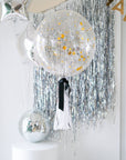 A large, clear balloon with a black bow and a white tassel, with 'Happy New Year' written on it, hanging indoors with silver tinsel background