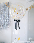 A large, clear balloon with a black bow and a white tassel, with 'Happy New Year' written on it, hanging indoors with party decorations around.
