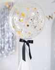 A large, clear balloon with a black bow and a white tassel, with 'Happy New Year' written on it, hanging indoors with party decorations around.