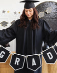 Graduate in cap and gown with 'GRAD' banner against a decorated wall.