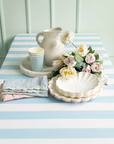 A pale blue stripe paper tablecloth with green stripes on a long table. Npakins, plates, flowers and vase set on top.