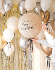 Cream round balloon with black bow and silver metallic tinsel. Text reads "3..2..1.. Happy new Year!" with gold tinsel and cream backdrop. Floating balloon with black ribbon bows surrounding.