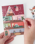 Hand holding a small house-shaped card with a sticker sheet featuring Christmas-themed stickers.