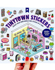 A hand with red nail polish holds a square sticker sheet featuring the 'Tinytown Stickers' design. The sticker sheet displays a colorful, illustrated bakery storefront with various themed stickers around it, including cupcakes, donuts, lollipops, and small characters.