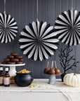 Black and white striped paper fan decorations hanging on a dark gray paneled wall above a styled Halloween dessert table with cookies, candy apples, a cauldron, and a white pumpkin.