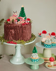 A scene with a large brown cake and small cupcakes next to it. All cakes h ave winter woodland themed cake toppers sitting on tnop of the cake.
