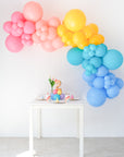 An indoor balloon garland featuring in a mix of colorful balloons in shades of pink, yellow, blue, and teal. Garland is above a small table.