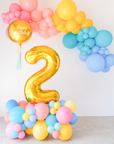 An indoor balloon garland featuring a large gold number 2 amidst a mix of colorful balloons in shades of pink, yellow, blue, and teal, with a 'Happy Birthday' message on the top large balloon.