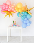 A balloon garland featuring a variety of colored balloons including pink, yellow, blue, and multicolored starburst and confetti balloons, displayed indoors next to a table with a decorative item.