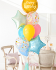 A balloon bouquet featuring a round gold orb balloon, with smaller multicolored balloons including pink, blue, and yellow, all adorned with confetti clear balloons and stars. The balloons are attached to a ribbon bow and tassel. Bouquet is in a dining room setting.