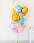 A balloon bouquet featuring two round gold orb balloon, with smaller multicolored balloons including pink, blue, and yellow, all adorned with confetti clear balloons and stars. The balloons are attached to a ribbon bow and tassel.