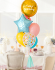 A balloon bouquet featuring a round gold orb balloon, with smaller multicolored balloons including pink, blue, and yellow, all adorned with confetti clear balloons and stars. The balloons are in a dining room