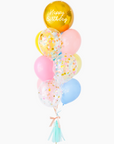 A balloon bouquet with a mix of solid colored balloons in blue, yellow, pink, and green, along with one metallic orb balloon with 'Happy Birthday' text, and a clear confetti balloon. There is a tassel and a ribbon bow on the arrangement.