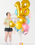 A balloon arrangement featuring a golden number '18' balloon with multicolored balloons tied to it, with the word 'Happy Birthday' printed on a gold balloon. A girl is standing next to it and look towards the bouquet.