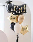 A balloon bouquet with black, gold, and silver balloons, including one that reads 'Grad' and star-shaped ones.