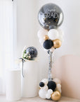 A balloon tower with a combination of silver, black, white, and gold balloons, with the top balloon saying 'Congrats GRAD'.