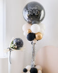 A balloon tower with a combination of silver, black, white, and gold balloons, with the top balloon saying 'Congrats GRAD'.