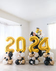 A person wearing a grad cap and gown is standing behind the balloon stacks arranged to form the numbers '2024' in gold with black and white balloons around them, displayed indoors.