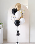 graduation balloon bouquet in black, white, cream. confetti-filled, and gold colors in front of an arch backdrop