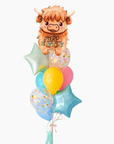 A balloon bouquet featuring a Highland cow balloon with the text 'heard it was your birthday' and several coordinating balloons including a yellow round balloon, pastel blue and pink balloons, and two light blue star-shaped balloons with confetti.