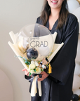 A person holding a mini bouquet with a large clear balloon in the center that has 'CONGRATS GRAD' written on it, surrounded by white and yellow faux florals and a small black bear figurine wearing a graduation cap.
