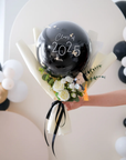 A hand holding a mini graduation bouquet with a large black balloon featuring 'Class 2025', wrapped with black and white paper, and decorated with white and yellow flowers and a small black bear figurine.