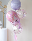 A bunch of balloons with 'Happy Mother's Day' written on one, in colors pink, purple, and clear, with some balloons having confetti inside.