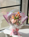 Bouquet of faux spring flowers in a transparent balloon with 'Happy Mother's Day' text, wrapped in lavender paper and ribbon by a window
