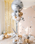 A round silver balloon with New Year text accompanied with smaller clusters of balloons underneath on a stand. Gold tinsel and cream backdrop in the background. Confetti accents are flying on the side.