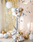 Confetti flying around from the popped round balloon accompanied with smaller clusters of balloons underneath on a stand. Gold tinsel and cream backdrop in the background.