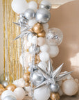 A balloon galrand in shiny silver, gold, white and creams with metallic tinsel and spikey balloon accents against a cream backdrop and gold tinsel curtain