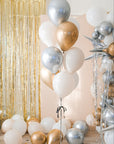 A larger bouquet of balloons in gold, silver, cream, and white colors with sparkly silver tinsel strings, displayed indoors with a glittery backdrop.