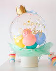 A clear bubble box with a crown on top, decorated with colorful balloons and confetti, and labeled 'Happy Birthday'. Cups near the box on the table.
