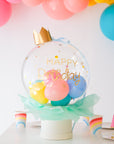 A clear bubble box with a crown on top, decorated with colorful balloons and confetti, and labeled 'Happy Birthday'.