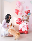 A Valentine's Day balloon present featuring a heart-shaped balloon with the name 'Olivia', a teddy bear holding balloons, and a corgi dog next to a woman kneeling down