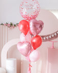 A balloongram featuring a large clear 'Be My Valentine' balloon, along with six latex balloons, two hearts, a tassel, and a bow for decoration, all in shades of pink and red.