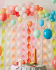 A string of crepe paper streamers in a rainbow color palette, with fringe-like ends, displayed against a white background with balloons arched over