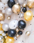 Assorted gold, silver, black, and white balloons scattered on the floor around a pink wall, with some balloons.