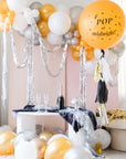 A balloon garland made of silver and gold balloons, set up in a room with a pink wall and a table with a bottle.