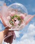 A clear balloon with 'Congratulations' text, filled with pink faux flowers, tied with a ribbon against a blue sky background.