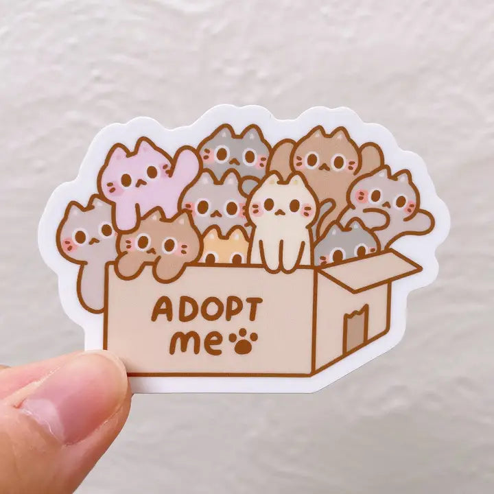 Sticker with cartoon cats in a box and 'ADOPT me' text on a plain background