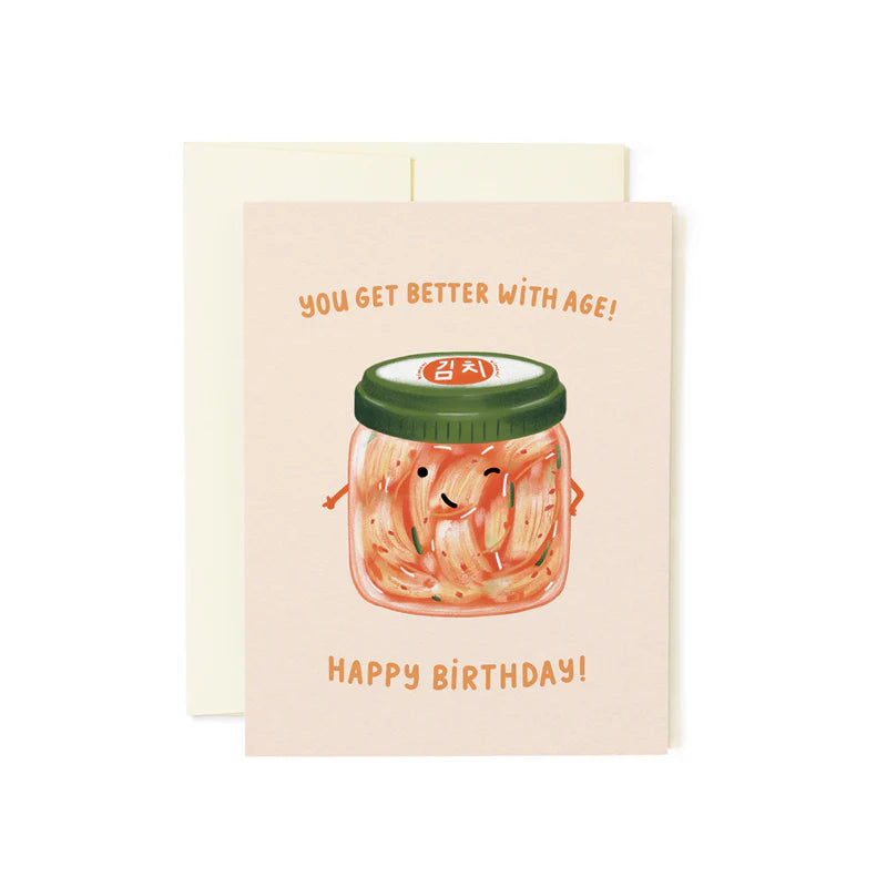 A greeting card with an illustration of a jar of kimchi and a lid, with the text 'YOU GET BETTER WITH AGE! HAPPY BIRTHDAY!' written on it. The card is pink, and there is a white envelope included.